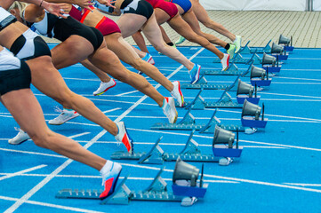 Start of the sprint. Running photo from the Track and Field event. Female professional athletes start from the sprint blocks on the blue tartan. Illustrative photography for athletic events