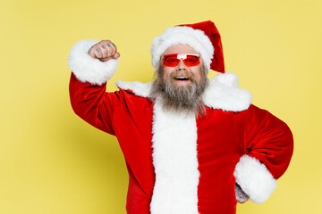 happy plus size santa claus in trendy sunglasses demonstrating power isolated on yellow.