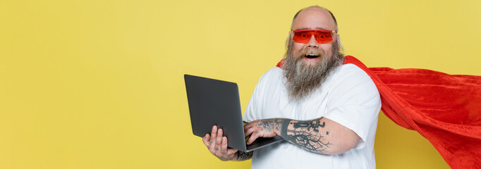 cheerful overweight man in red sunglasses and superhero cloak using laptop isolated on yellow,...