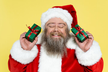 pleased and chubby santa claus with closed eyes holding gift boxes isolated on yellow.
