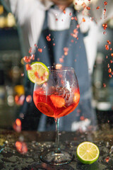 Bartender pours cocktail into glass of ice painted with lime, spray flies