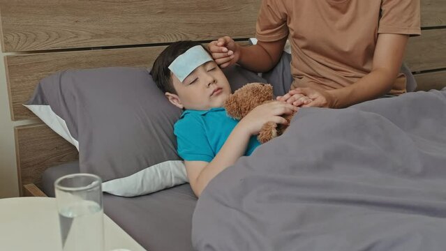 Tilt up shot of sick Asian boy with cooling pad on forehead sleeping with bear toy in bed at home while careful mother holding his hand