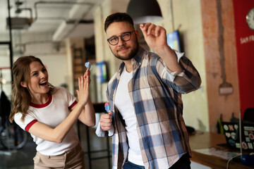 Young colleagues taking break after work. Happy young businessman and businesswoman playing darts...