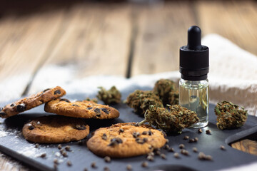 Homemade chocolate chip cookies with cbd oil, surrounded by dry cannabis buds, marijuana seeds and a glass bottle with a dropper of cbd oil.  On a stone board, on a wooden background