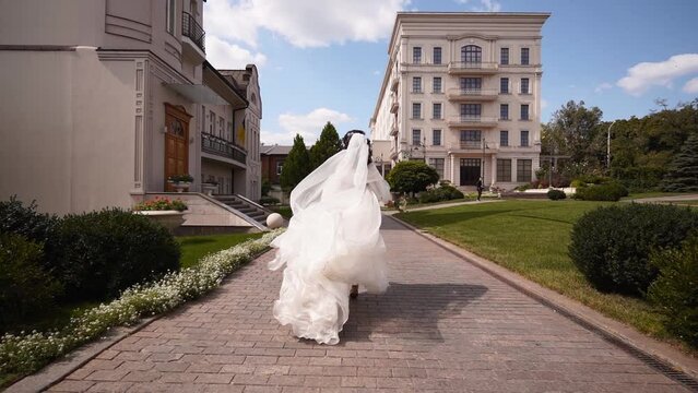 Pretty bride in long airy veil, elegant white dress runs away from camera on wedding day. Playful attractive woman running away turning, looking to viewer. Follow me concept. Fabric fluttering in air.