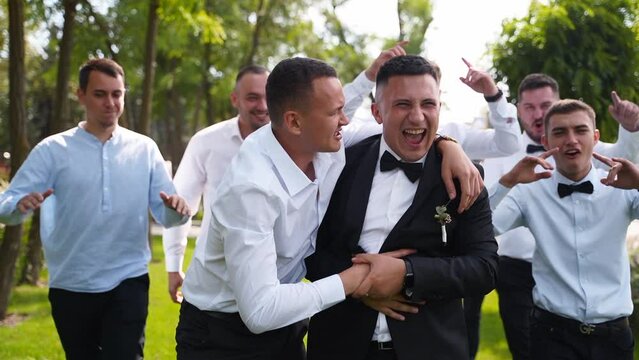 Groom flexing and having fun walking with groomsmen on wedding day. Happy classy man in black suit, bow tie and boutonniere hanging out partying with friends and cheering posing at camera in a park.