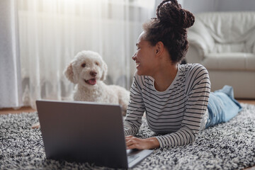 Cheerful smiling female typing on laptop computer resting at cozy room on carpet with pet