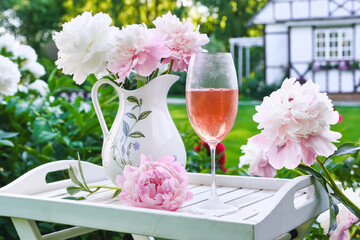 Still life with wine glass with rose wine on a wooden tray with a bouquet of peonies and jug on a blurred background in the garden. Concept of celebration party outdoors in a summer day.