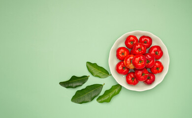 Ripe red acerola cherries in a white dish and green leaves scattered on a green background