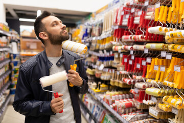a man with different rollers for painting walls looks thoughtfully at a rack with brushes