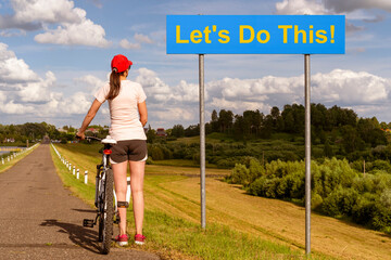 Young woman cyclist reads text Let's Do This! written on a highway road sign