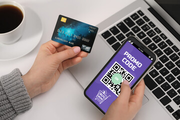 Woman holding credit card and smartphone with activated promo code in online shopping app at white table, closeup