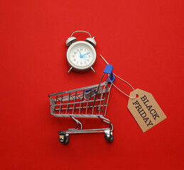 Shopping cart, tag with phrase BLACK FRIDAY and alarm clock on red background, flat lay