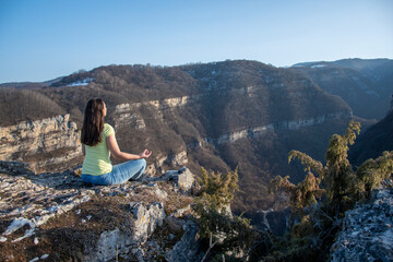 A girl sits on the edge of a cliff in a lotus position, meditates in blue jeans and a yellow T-shirt, view from the back