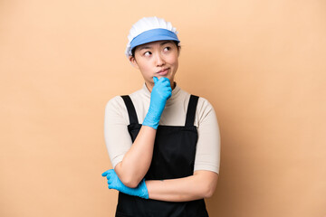 Young Chinese Fishmonger woman wearing an apron and holding a raw fish isolated on pink background having doubts