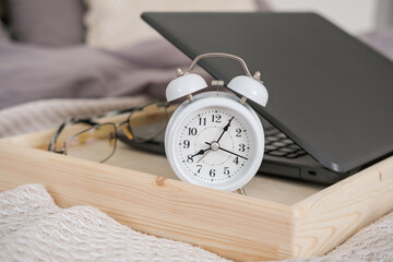 Naklejka premium Laptop, alarm clock and glasses on a tray on the bed in the bedroom. Time to work. The concept of work at home and distance learning.