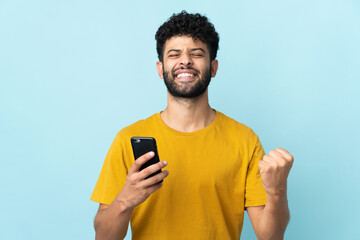 Young Moroccan man isolated on blue background using mobile phone and doing victory gesture
