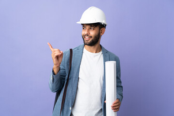 Young architect Moroccan man with helmet and holding blueprints over isolated background intending...