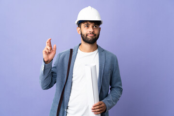 Young architect Moroccan man with helmet and holding blueprints over isolated background with fingers crossing and wishing the best