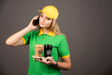 Young deliverywoman with cups of coffee speaking on smartphone