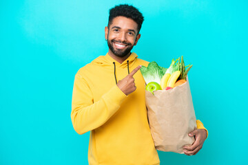 Young Brazilian man taking a bag of takeaway food isolated on blue background pointing to the side to present a product