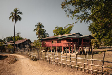 Don Det, Laos - January 18th, 2020 : Colorful red and green lao wooden house on stilts with palm trees in the background and a bamboo fence and sandy pathway in the foreground. - obrazy, fototapety, plakaty