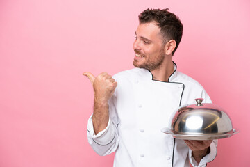 Young Brazilian chef with tray isolated on pink background pointing to the side to present a product