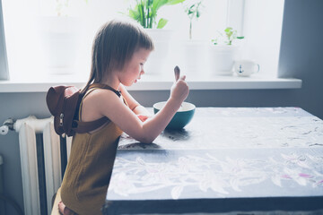 cute toddler child with backpack eats at table by window