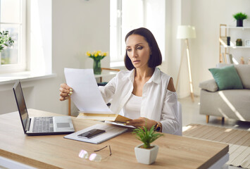 Serious worried mature woman reading utility bill sitting at work desk with laptop computer. Entrepreneur receives debt notice. Middle aged businesswoman gets paper letter reminding of overdue payment