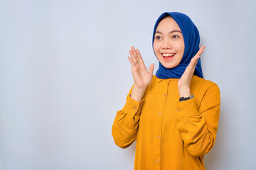 Shocked young Asian Muslim woman dressed in orange shirt looking at copy space and raising hands with open mouth, reacting to sale offer isolated over white background