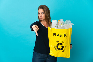 Middle age Brazilian woman holding a bag full of plastic bottles to recycle on blue background showing thumb down with negative expression