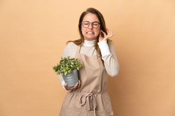 Middle age gardener woman holding a plant isolated on beige background frustrated and covering ears