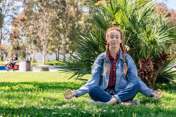 Naklejka na ściany i meble A smiling woman with pigtails sits with her eyes closed in a lotus position on a green lawn near a palm tree. Meditation in the southern solar park