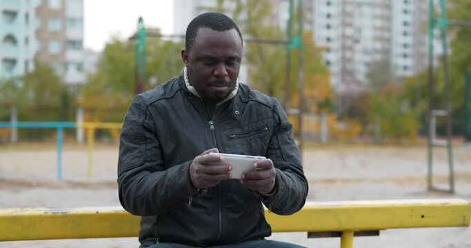 African american man sits at sport ground and plays the game on smartphone