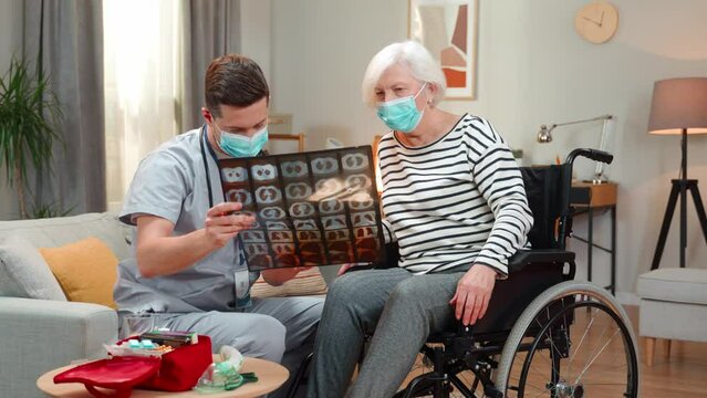 Caucasian young professioanal doctor in medical mask showing X-ray and talking to senior lady on wheelchair at home. Retirement, covid pandemic, physical disability. Healthcare concept