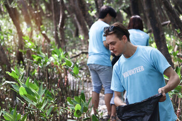 Group of volunteers in blue t-shirt hold garbage bag, ecology people cleaning and picking up trash to reduce land pollution environmental problem at mangrove forest, world environment day concept.