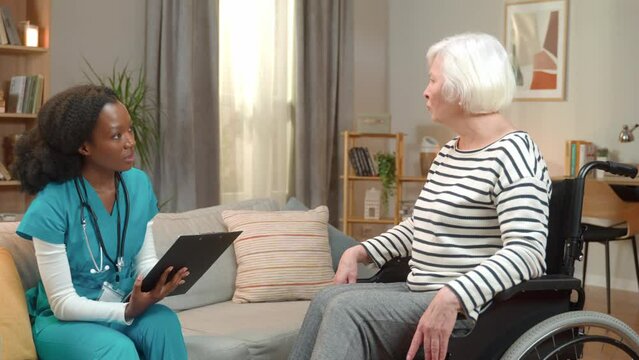 Portrait of cheerful old pretty disabled woman in wheelchair laughing while speaking with young female nurse at home visit in living room. Healthcare assistance, medicine, nursing, social help