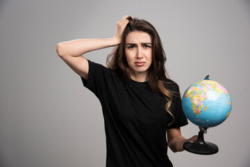 Young woman holding globe on gray background while covering her mouth