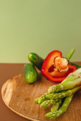 Vegetables lie on a wooden board: tomatoes, asparagus, cucumbers, red bell peppers. brown, light green background. place for text.