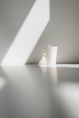 A white tall porcelain coffee cup on a white table with sun hitting the wall behind creating a nice morning atmosphere Nr.2