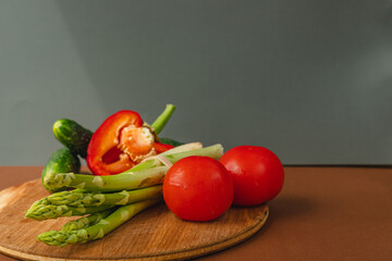 Vegetables lie on a wooden board: tomatoes, asparagus, cucumbers, red bell peppers. brown, dark gray background. place for text.