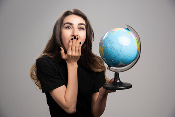 Young woman holding globe on gray background while covering her mouth