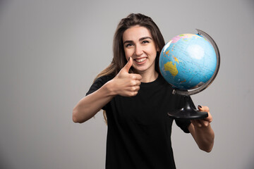 Fototapeta premium Happy woman with globe making thumbs up on gray background