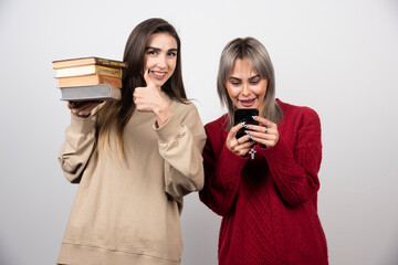 One girl holding a stack of books another girl looking at phone