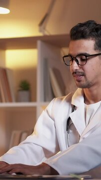 Vertical video. Working male doctor. Online communication. Digital technology. Handsome positive man typing laptop sitting work desk light room interior.