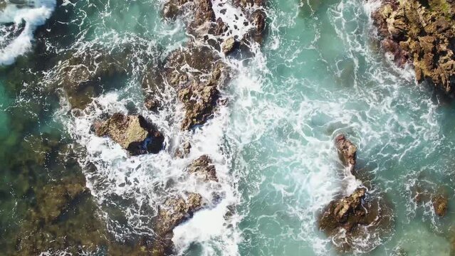 4K aerial shot from a drone, the purest turquoise waters break on the stone shore and rocks and foam. Surf splashes are scattered in slow motion