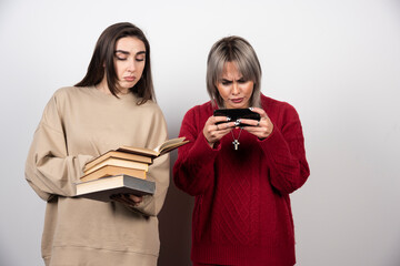 One girl holding a stack of books another girl looking at phone