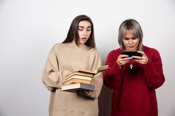 One girl holding a stack of books another girl looking at phone
