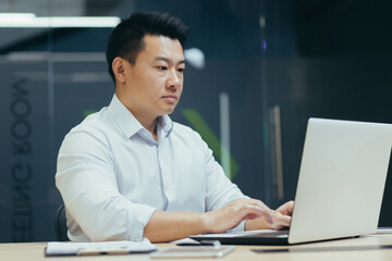 Serious and confident asian boss working in modern office, businessman typing on laptop.