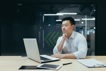 Fototapeta na wymiar Pensive asian businessman working in modern office, man thinking about decision, experienced director with laptop.